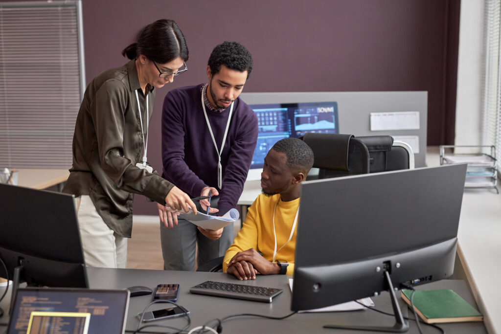 Three cybersecurity professionals huddle up around a computer station to review the results of their CMMC mock audit.